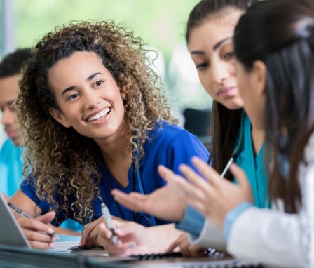 A smiling young woman listens attentively during a discussion with her classmate and professor. She is sitting in the classroom and working on a medical training assignment with her classmate. Image credit: SDI Productions / E+ / Getty Images