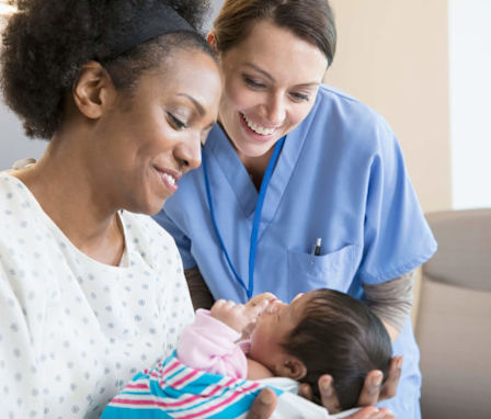 A mid-adult Caucasian nurse midwife is checking in with an African-American mother and her newborn child. The mother is lying in a hospital bed, swaddling her baby in her arms and smiling. The midwife is wearing light blue scrubs. She is standing over the mother and baby and smiling while she does her health check-in with them.