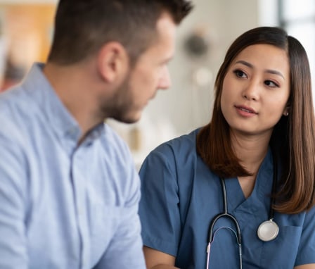 A young female Asian-American nurse and a young male Caucasian patient are chatting inside a clinical office. The nurse is listening to the man while holding a tablet computer she uses to take notes. She has long brown hair and is wearing blue scrubs and a stethoscope.