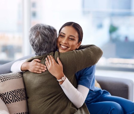 A young female nurse smiles while being hugged by a senior female patient. They are sitting down on a couch inside a hospital waiting room.
