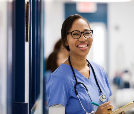 A Black mid-adult female nurse looks up from her patient chart to give the camera a big smile.