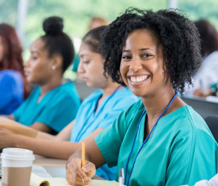 A smiling mid-adult African American nursing student is sitting in class with her colleagues. She has short, curly black hair, and is wearing teal green scrubs. Her laptop and notepad are open so she is ready to take notes during class. She is looking directly at the camera.
