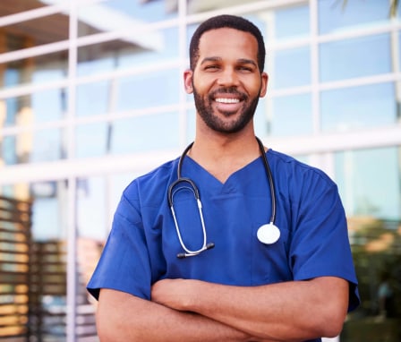 A young black male nurse standing outside a hospital smiling at the camera.