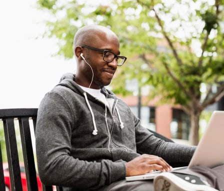 A Black male student sitting in a park bench working on his laptop.