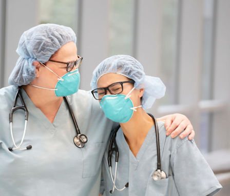 Two female, health care providers laugh while wearing light green medical scrubs, stethoscopes and N95 masks in an indoor hallway.